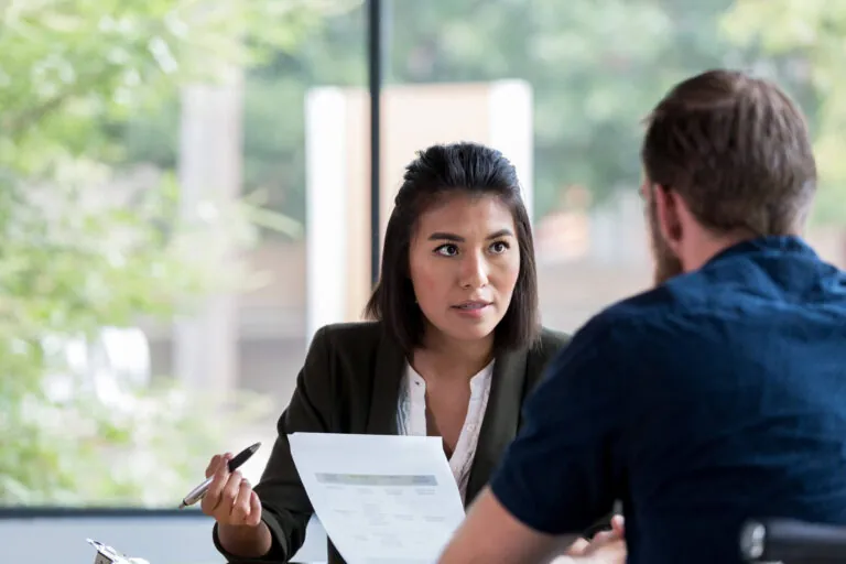 Serious Hispanic businesswoman shows a document to a male business associate. The business associate attentively listens to the businesswoman.
